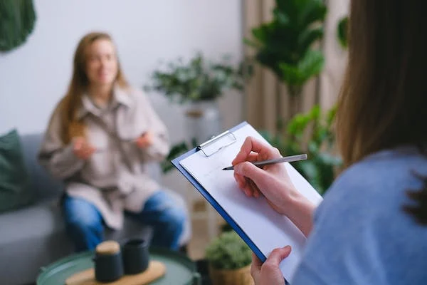 A person writing on paper in front of another person.